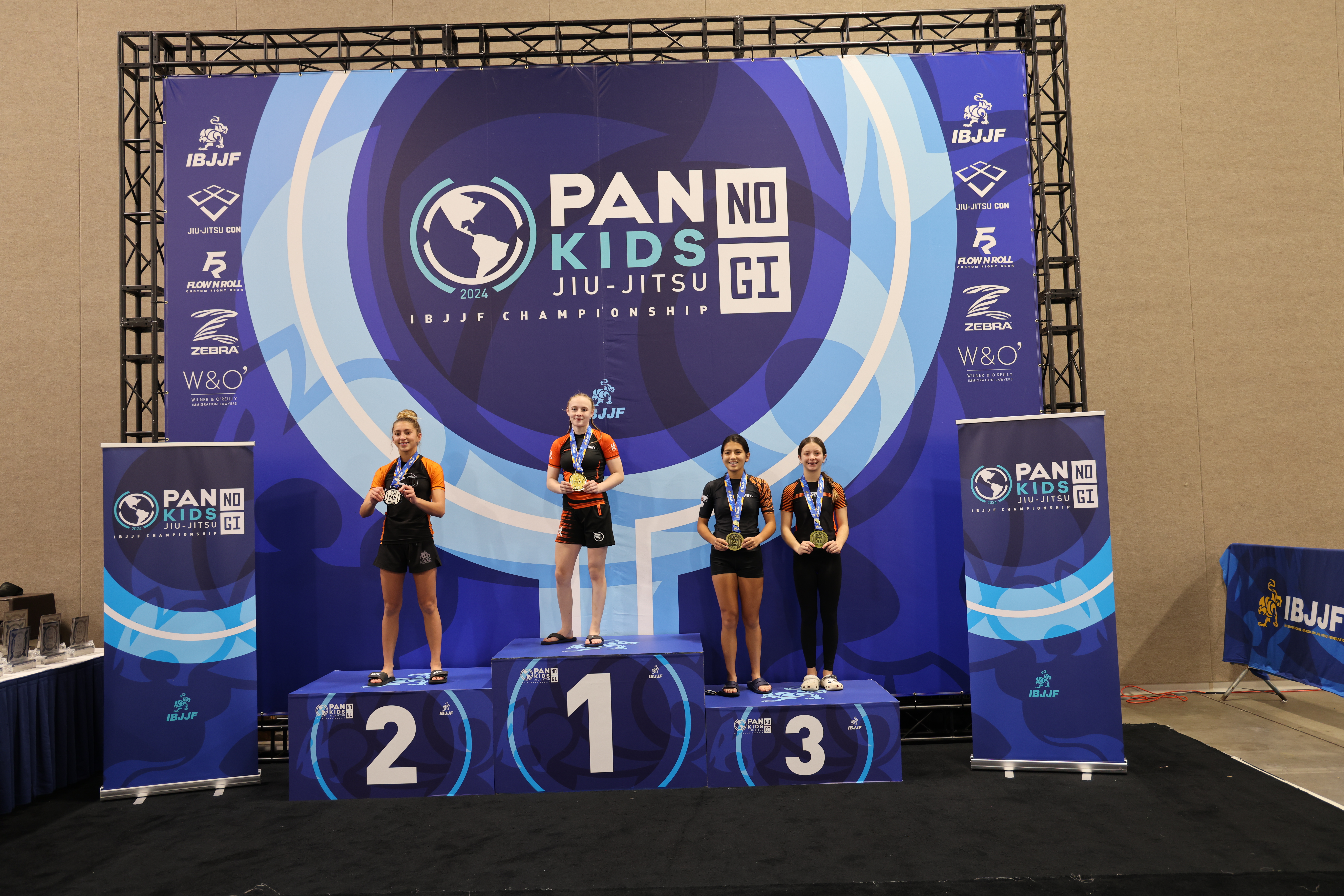 Podiumsfotografie von der Pan Kids IBJJF No-Gi Championship 2024. Die oberste Stufe des Podiums ist von der Divisionssiegerin besetzt. Im Hintergrund ist das Event-Branding sichtbar: PAN KIDS NO-GI IBJJF CHAMPIONSHIP.